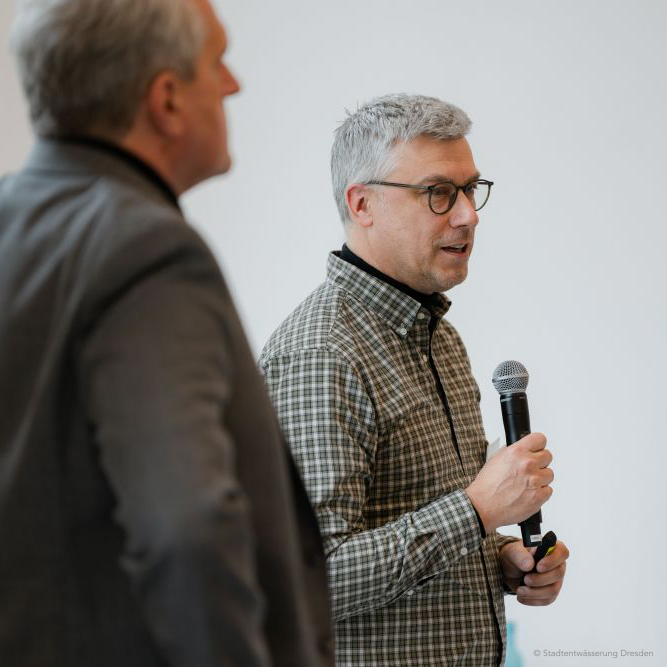 Robert Storch spricht mit Mikrofon im Handgriff; graue Haare und schwarze Brille sowie ein kariertes Hemd prägen den Moment.