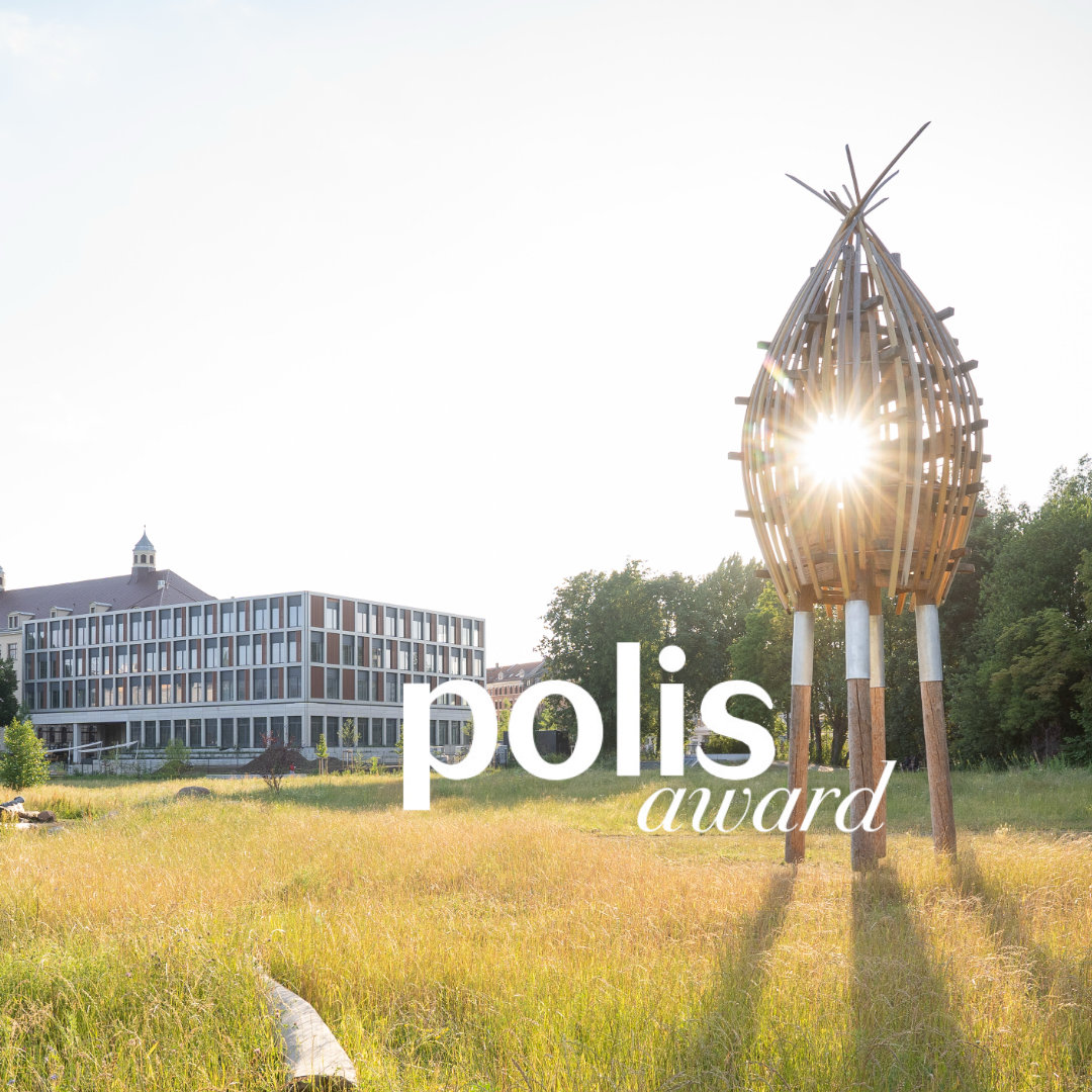 Helle Wiese mit großer Holzkonstruktion im Vordergrund, moderne Gebäudefassade im Hintergrund und der Text „polis award“ in Weiß über der Szene